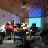 AI: Classroom with white ceiling, wooden panels, recessed lighting; blue screen showing presentation; tables with pairs, one person holding Hello sign (AI-generated alt-text)