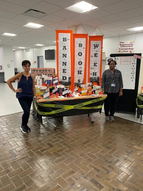 Marta Weigand and Deborrah Tomengo stand on either side of their Banned Books Week display, A number of books sit out on a table that is wreathed in black cloth, tinged with paper fire, and wrapped in yellow caution tape. Red banners reading "banned books week" hang from the ceiling above. 