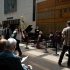 AI: Outdoor scene at San Francisco Conservatory: brick sidewalk, black piano with pianist, guitarist, saxophonist, audience on folding chairs listening (AI-generated alt-text)