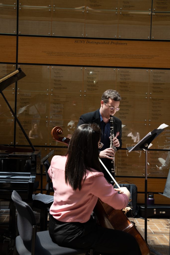 AI: Man and woman playing violins on black chairs in UW‑Milwaukee lobby with dark wood wall, gold lettering, window (AI-generated alt-text)