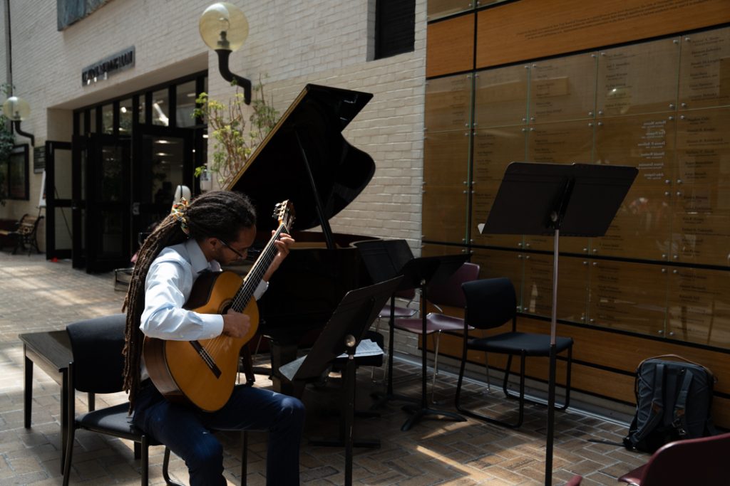 AI: Man with dreadlocks plays light brown guitar on chair outside brick music store; black music stands, backpack on nearby chair (AI-generated alt-text)