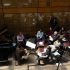 AI: Seven people, six seated listening, one standing near piano, wooden walls, window left, piano with stand, red stool foreground (AI-generated alt-text)