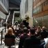 AI: Outdoor courtyard day event with people sitting and standing, man in suit on stage holding microphone, modern glass buildings (AI-generated alt-text)