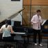 AI: Three musicians perform: pianist at black piano, violinist with bow, and violinist, in front of wooden façade and brick wall (AI-generated alt-text)