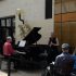AI: Trio performing on sidewalk: black piano, man in red playing guitar, woman in dress playing saxophone, rope barrier, yellow building (AI-generated alt-text)