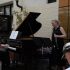 AI: Trio on sidewalk outside JAZZ SAW: red‑shirt pianist, black‑dress sax player, blue‑shirt dancer with mic, brick wall, gold sign (AI-generated alt-text)