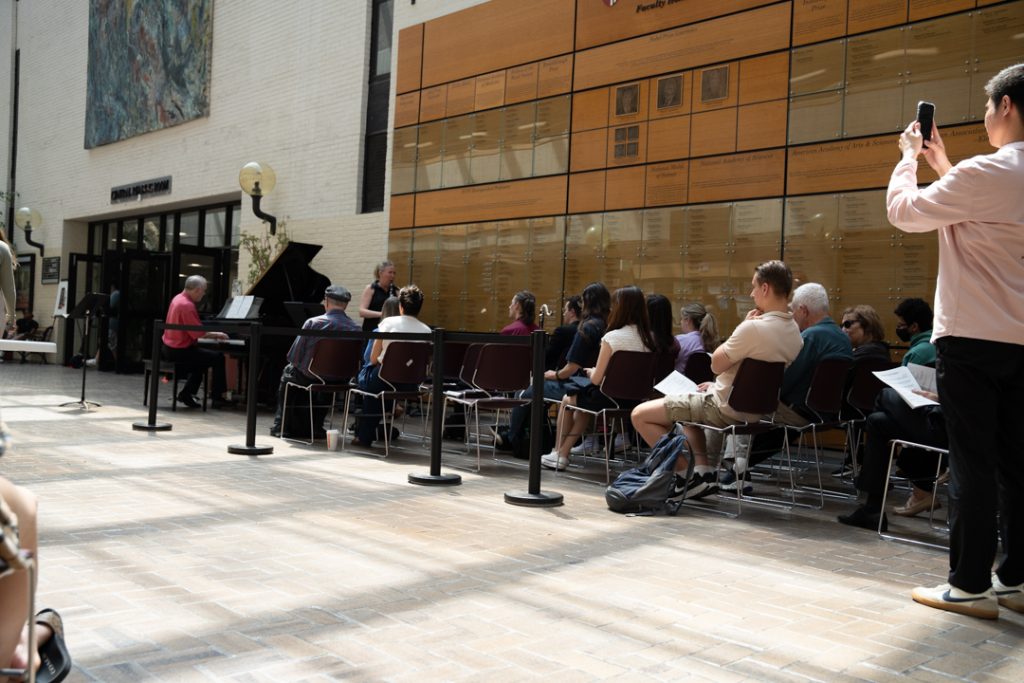 AI: Three people seated by a glass storefront with BANQUETING sign: red guitarist, blue listener, pink photographer; brick floor, passerby walking (AI-generated alt-text)