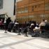 AI: Three people seated by a glass storefront with BANQUETING sign: red guitarist, blue listener, pink photographer; brick floor, passerby walking (AI-generated alt-text)