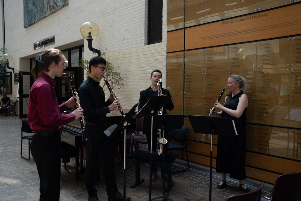 AI: Four musicians on brick sidewalk before glass building: two saxophonists, bearded player, violinist, sign PARKER HOUSE BOSTON (AI-generated alt-text)