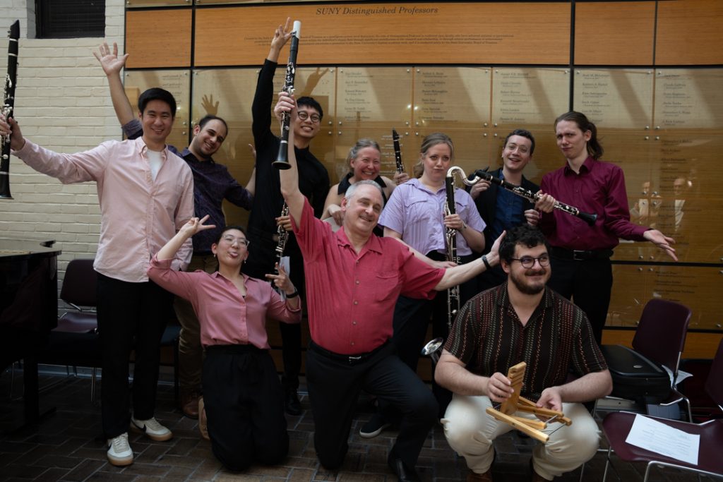 AI: Eight people kneeling in two rows before brick wall with window; back row holds black instruments, front row claps (AI-generated alt-text)