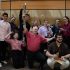 AI: Eight people kneeling in two rows before brick wall with window; back row holds black instruments, front row claps (AI-generated alt-text)