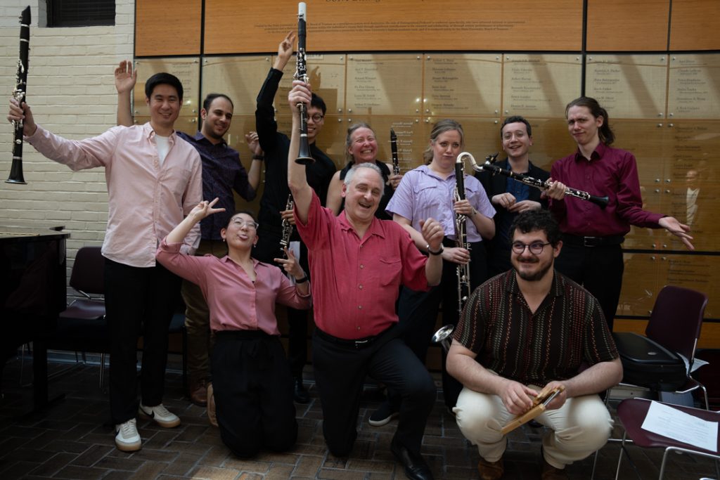AI: Nine musicians stand before a brick wall, holding trumpet, guitar, violin and other instruments, facing camera (AI-generated alt-text)