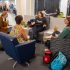 AI: Woman in black shirt with SKY logo, pink floral tank, sweater on chairs; coffee table with cooler, cup, plant; cat nearby (AI-generated alt-text)