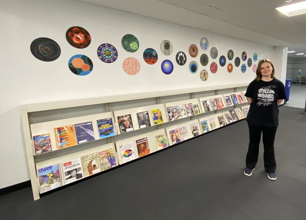 Picture of a student wearing a black shirt and black pants in front of a multicolored circles on a wall.