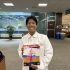 AI: Young man with glasses in lab coat holds white book with pink-purple cover, red FIRST AID badge, office with windows (AI-generated alt-text)