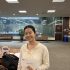 AI: Young woman in library holding paper saying I'm sorry, smiling; light blouse, wooden floor, window behind, bench and trash can (AI-generated alt-text)