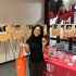 AI: Young woman smiling holds red Staples bag beside table orange‑black balloons, signs, brochures and Dance for a Cure flyers, indoor (AI-generated alt-text)