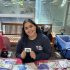 AI: Young girl wearing Who's got milk? shirt smiles, holds merchandise at table with bags, stickers, park view, bench, trash can (AI-generated alt-text)