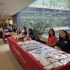 AI: Lively store display with red table covered in colorful gift bags, three people arranging them, warm lighting, window background (AI-generated alt-text)