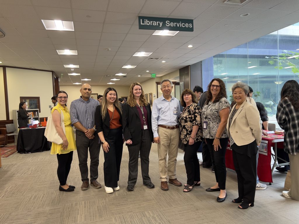 AI: People stand under green Library Services sign, counter with computer behind; one person in yellow jacket (AI-generated alt-text)