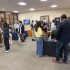 AI: Eight people in an office; four stand by a monitor desk, three sit on blue and gray chairs, woman approaches (AI-generated alt-text)