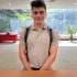 AI: Young man in gray polo and backpack smiling at camera beside wooden table, monitor, keyboard, window with trees, plant (AI-generated alt-text)