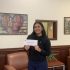 AI: woman holding card with hello written, wearing black sweater, standing by bookshelf and two paintings (AI-generated alt-text)