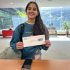 AI: Young woman in blue sweater holds torn card reading Kia Sorento in library, backpack, people behind, window with natural light (AI-generated alt-text)