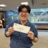 AI: Young man in blue shirt holding Super Bowl business card, smiling with glasses, standing in office with wall and desks (AI-generated alt-text)