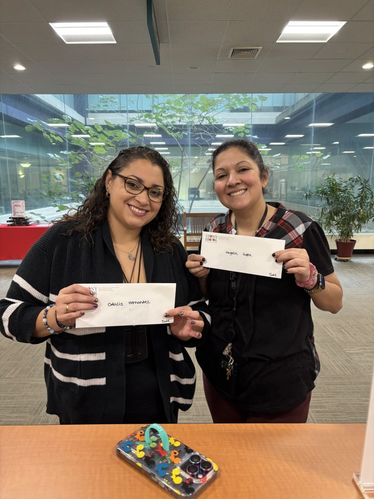 AI: Two women in office by window, smiling, holding white We have love signs; left black cardigan, right red shirt (AI-generated alt-text)