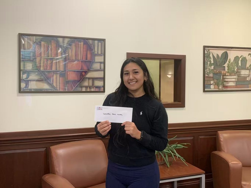 AI: Young woman holding a New York card in front of a bookshelf with heart design, two paintings on wall (AI-generated alt-text)