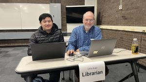 Shows two smiling people sitting behind a desk with laptops out