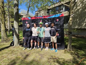 Picture of five people posing in front of a black backdrop with Stony Brook University logo on it. They are standing outside surrounded by trees.